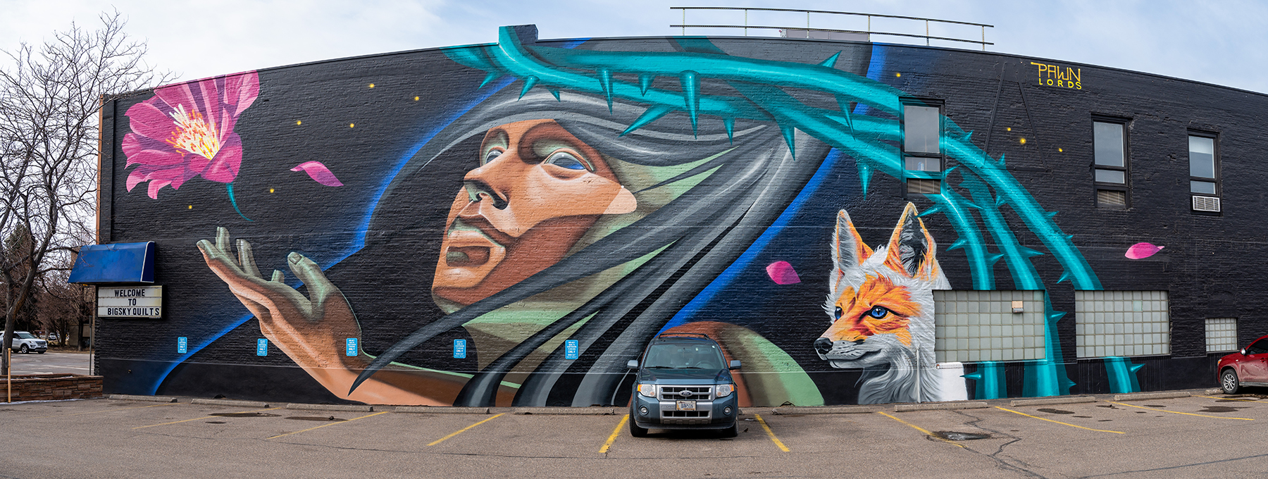 Image of mural on building wall featuring human face, thorny vines, a fox, and bitterroot flower